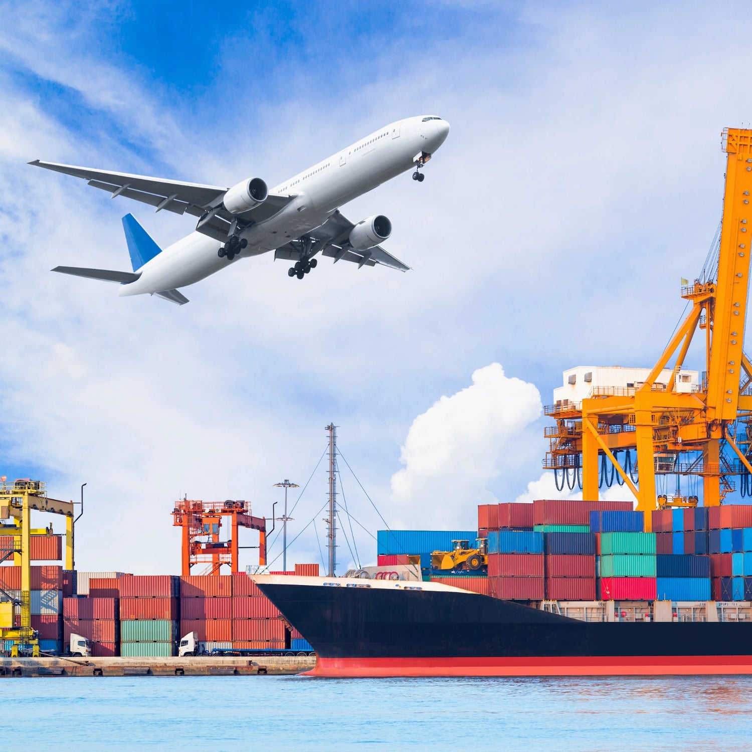 Airplane flying over a container ship and port with cranes against a blue sky.