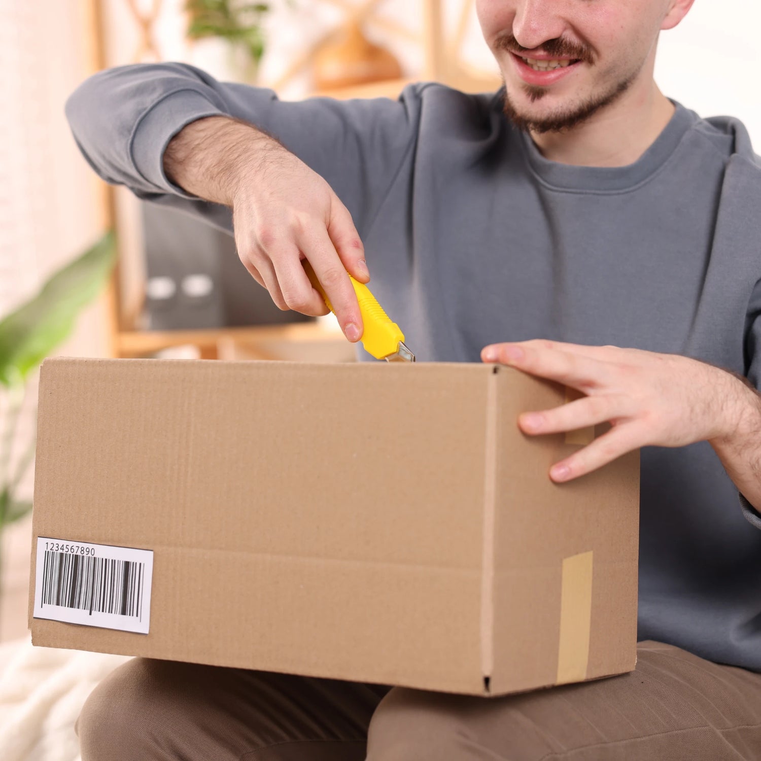 Man opening a cardboard box with a yellow tool, sitting on a couch.
