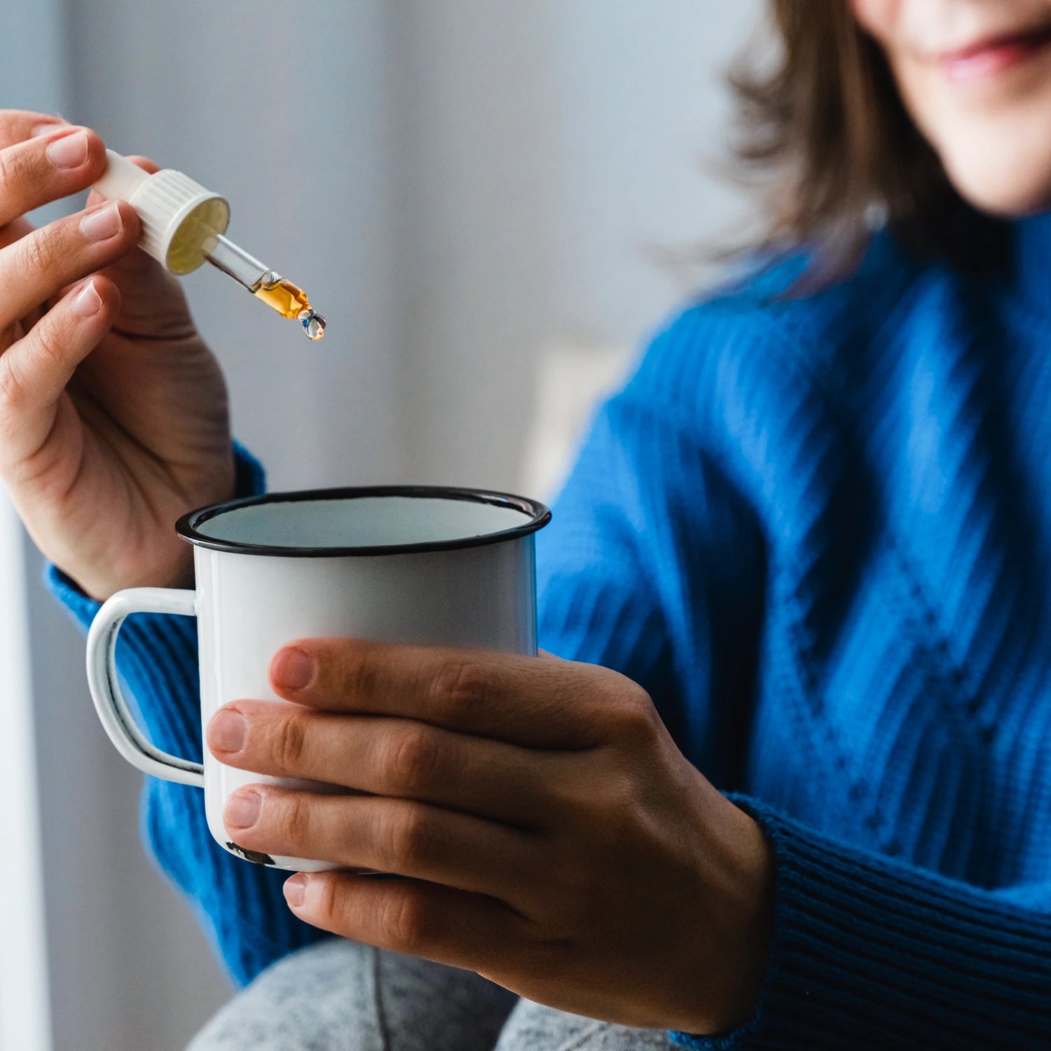 Person holding a white mug and a dropper with CBD oil, against a neutral background.